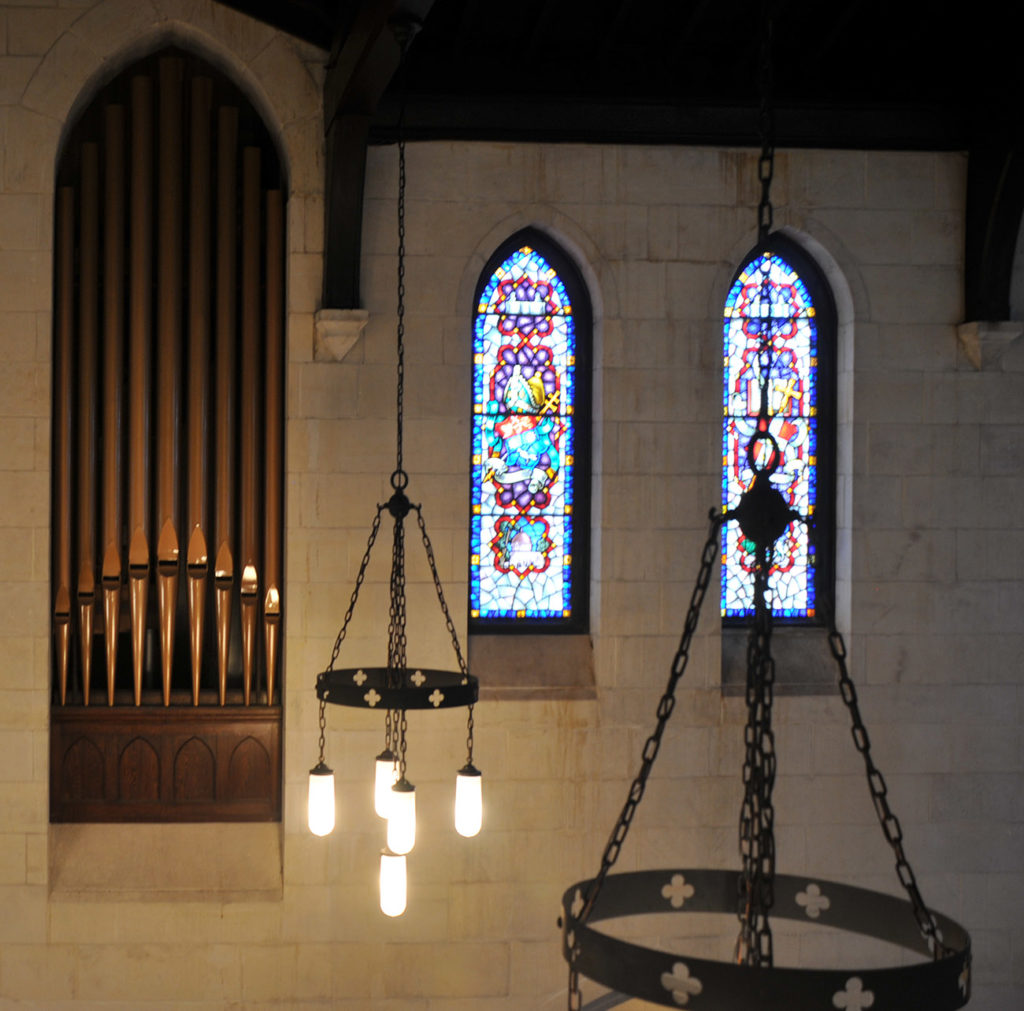Church interior near the ceiling. Two stained glass windows in the background on the right. Organ pipes in the walls on left. In the foreground are two metal chandeliers hanging from the ceiling.